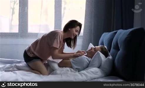 Young couple in love relaxing together in the bed after waking up in the morning. Playful girl riding on sleepy guy, tickling him to wake up. Affectionate couple embracing and kissing in bedroom.