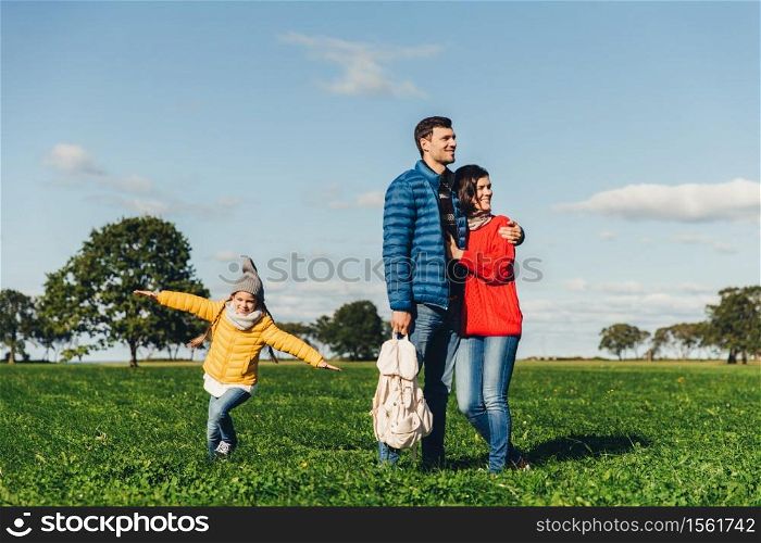 Young couple in love stand on green grass, embrace and look pensively. Playful small child enjoys freedom, runs on field, plays near her parents, has good mood and recreation. Peope and nature