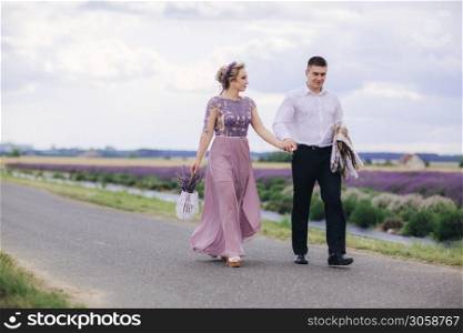 Young couple in love walking near a lavender field on summer day. girl in a luxurious purple dress and with basket of flowers. Young couple in love walking near a lavender field on summer day. girl in a luxurious purple dress and with basket of flowers.