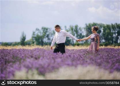 Young couple in love walks holding hands in a lavender field on summer day. girl in a luxurious purple dress.. Young couple in love walks holding hands in a lavender field on summer day. girl in a luxurious purple dress