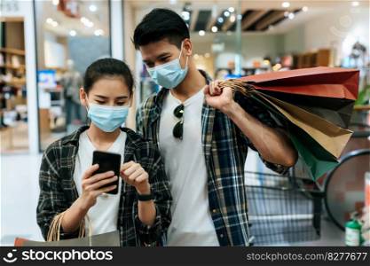 Young couple in protection mask holding multiple paper shopping bag and use smartphone while walking in the corridor of large shopping mall, New normal lifestyle and shopping concept