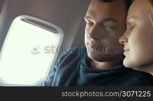 Young couple in the plane. Man looking out the illuminator and then trying to fall on sleep leaning on girlfriends head