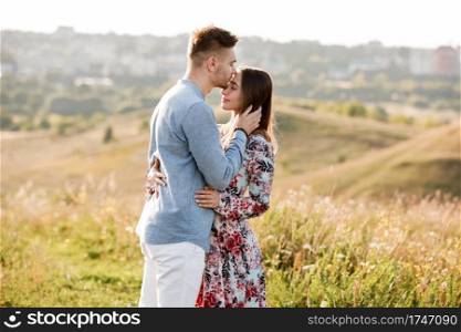Young couple is hugging and walking in summer field with grass on the background town. Man and woman. Concept of lovely family. Young couple is hugging and walking in summer field with grass on the background town. Man and woman. Concept of lovely family.
