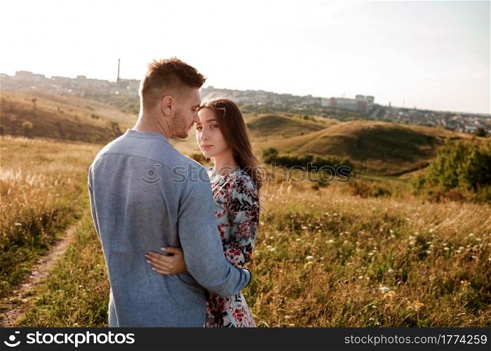 Young couple is hugging and walking in summer field with grass on the background town. Man and woman. Concept of lovely family. Young couple is hugging and walking in summer field with grass on the background town. Man and woman. Concept of lovely family.