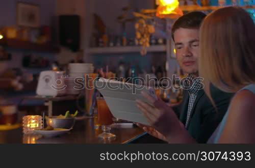 Young couple is sitting by the bar counter with tablet and drinking beverages from the glasses.