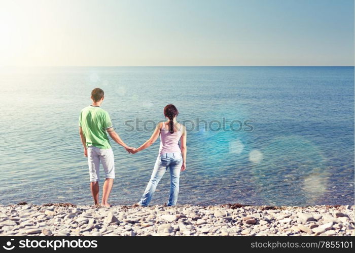 Young couple is standing near water, holding each others arms and looking over the horizon, view from behind, lens flares