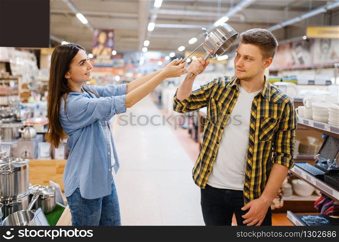Young couple jokes with ladle and pan in houseware store. Man and woman buying home goods in market, family in kitchenware supply shop. Couple jokes with ladle and pan in houseware store