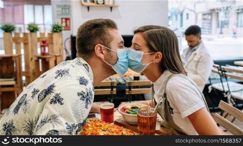 Young couple kissing with mask sitting in a restaurant. Couple kissing with mask sitting in a restaurant