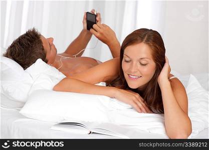Young couple lying down in bed, woman reading book, man listening to music