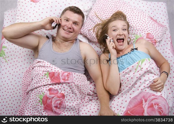 Young couple lying in bed. Everyone in my hand phone. Pair with gladness and joy of talking on the phone. Photo made at the top
