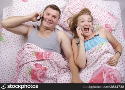 Young couple lying in bed. Everyone in my hand phone. Pair with gladness and joy of talking on the phone. Photo made at the top