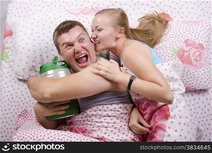 Young couple lying in bed. Husband with a barrel of beer, wife trying to take barrel bites her husband&#39;s ear