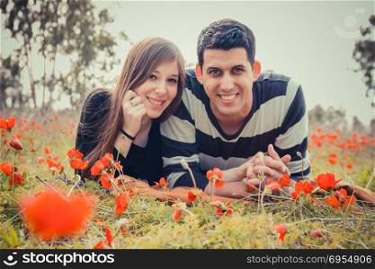 Young couple lying on the grass in a field of red poppies and smiling at the camera.
