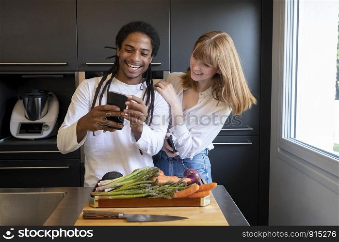 young couple of different ethnic groups with mobile phone in a kitchen with vegetable board. concept of healthy living.