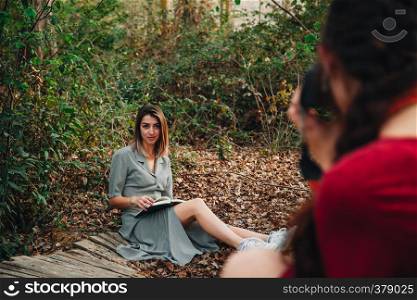 Young couple of women wearing dresses reading a book and taking photos with old camera in the forest