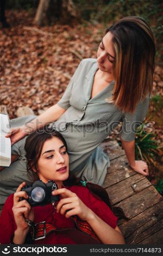 Young couple of women wearing dresses reading a book and taking photos with old camera in the forest