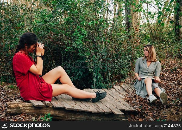 Young couple of women wearing dresses reading a book and taking photos with old camera in the forest with their horse.