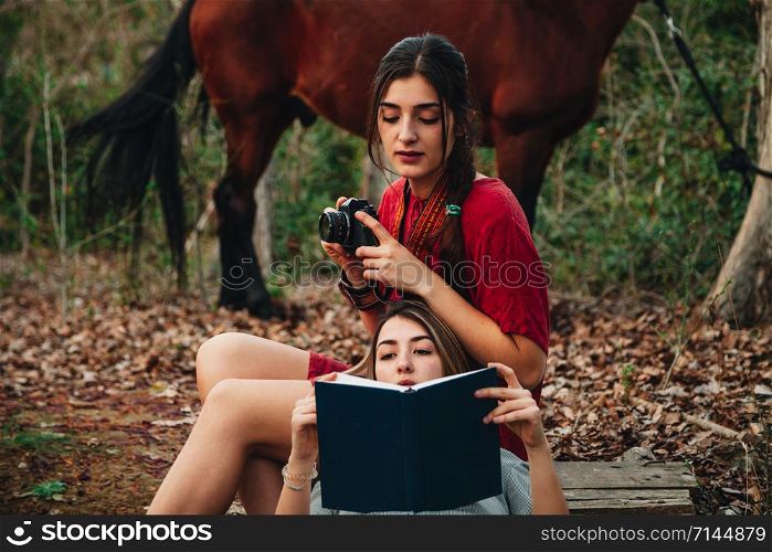 Young couple of women wearing dresses reading a book and taking photos with old camera in the forest with their horse.