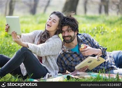Young couple on picnic blanket taking digital tablet selfie in woodland