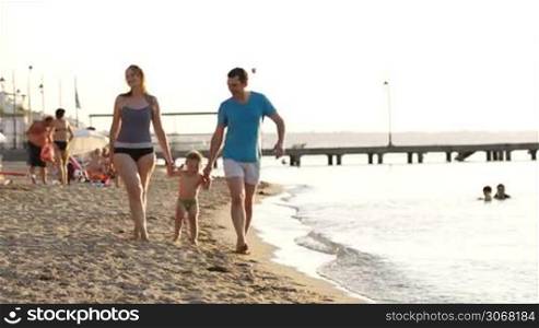 Young couple playing with their son at the seaside swinging him up in the air between them by his arms as they walk along the beach in their swimsuits