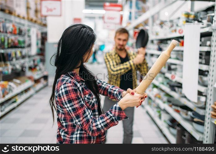 Young couple plays with frying pan and rolling pin in supermarket. Male and female customers on family shopping. Man and woman purchasing goods for the house. Couple with pan and rolling pin in supermarket