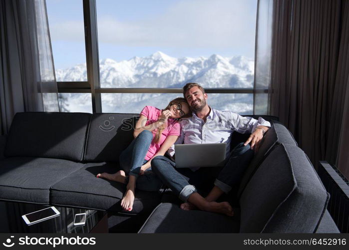 Young couple relaxing at home using laptop computers reading in the living room near the window on the sofa couch.