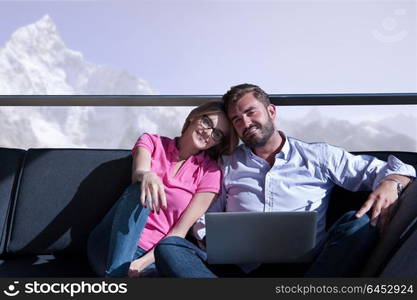 Young couple relaxing at home using laptop computers reading in the living room near the window on the sofa couch.