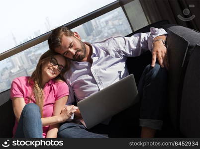 Young couple relaxing at home using laptop computers reading in the living room near the window on the sofa couch.