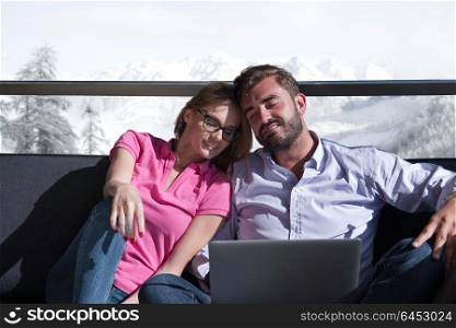 Young couple relaxing at home using laptop computers reading in the living room near the window on the sofa couch.