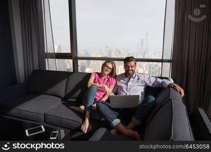 Young couple relaxing at home using laptop computers reading in the living room near the window on the sofa couch.