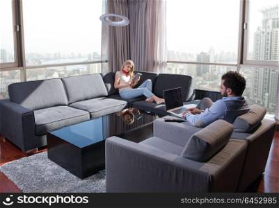 Young couple relaxing at home using tablet and laptop computers reading near the window on the sofa couch.