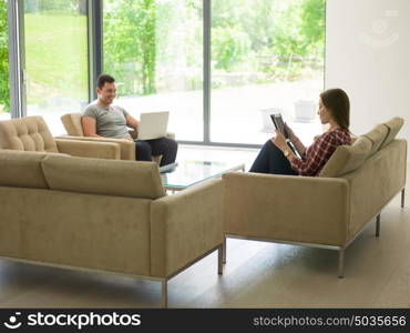 Young couple relaxing at luxurious home with tablet and laptop computers reading in the living room on the sofa couch.