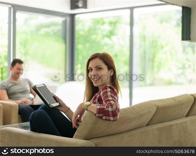 Young couple relaxing at luxurious home with tablet and laptop computers reading in the living room on the sofa couch.