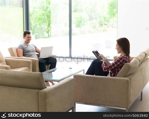 Young couple relaxing at luxurious home with tablet and laptop computers reading in the living room on the sofa couch.