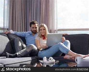 Young couple relaxing at luxury home using laptop computer reading in the living room on the sofa couch.