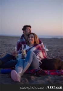 Young Couple Relaxing By The Fire, Drinking A Beer Or A Drink From The Bottle.. Loving Young Couple Sitting On The Beach beside Campfire drinkin