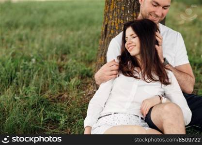 Young couple relaxing under tree in park on sunny day. Happy couple in love spend time outdoors together. Handsome man and pretty girl sitting on green grass leaning against tree.. Young couple relaxing under tree in park on sunny day. Happy couple in love spend time outdoors together. Handsome man and pretty girl sitting on green grass leaning against tree