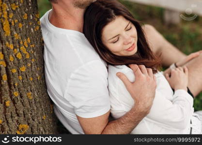 Young couple relaxing under tree in park on sunny day. Happy couple in love spend time outdoors together. Handsome man and pretty girl sitting on green grass leaning against tree.. Young couple relaxing under tree in park on sunny day. Happy couple in love spend time outdoors together. Handsome man and pretty girl sitting on green grass leaning against tree