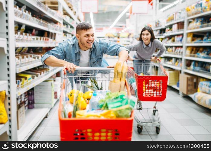Young couple ride on trolleys in supermarket, funny family shopping. Customers in shop, buyers in market