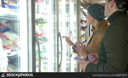 Young couple shopping for food in the supermarket. Woman taking product from the fridge. Man using pad to check shopping list