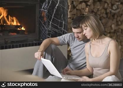 Young couple sit with laptop beside open fire