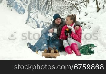 Young couple sitting by bonfire and drinking coffee in winter forest