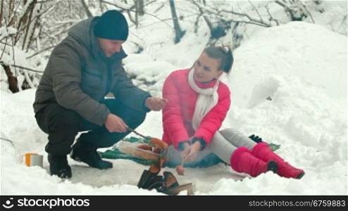 Young couple sitting by bonfire and roasting sausages in winter forest