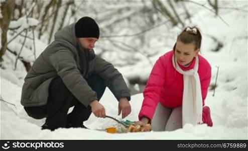 Young couple sitting by bonfire and roasting sausages in winter forest