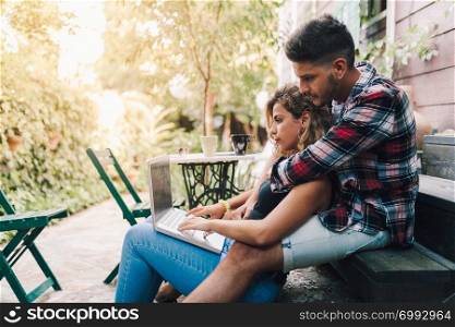 Young couple sitting embraced in the steps of their wooden house with a laptop