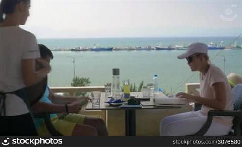 Young couple sitting in a cafe, the waitress takes the order from the woman, the man studying the menu. In the background a beautiful view of the sea