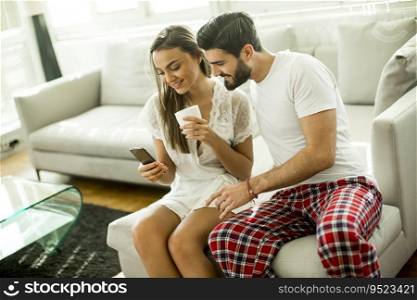 Young couple sitting on a sofa after waking up and looking at the phone, woman drinking coffee