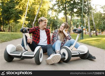 Young couple sitting on road with gyro board in summer park. Outdoor recreation with electric gyroboard. Eco transport, electrical gyroscope vehicle. Young couple sitting on road with gyro board