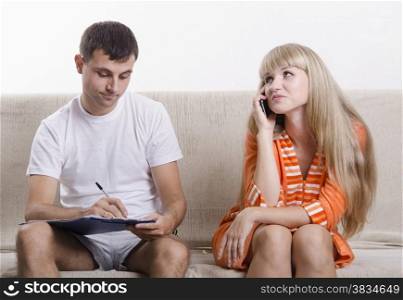 Young couple sitting on the couch. Girl on the phone, the guy sitting with the directory and writes a note of the telephone conversation. The pair dressed home