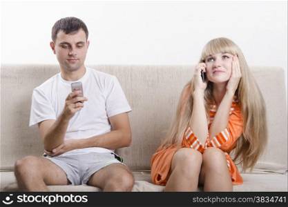 Young couple sitting on the couch. Girl on the phone. The guy writes SMS. The pair dressed home